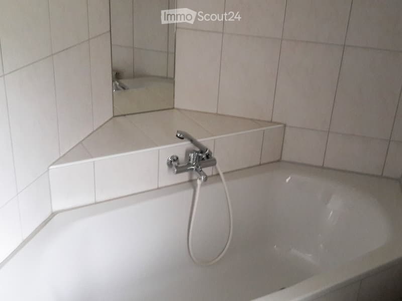Bathroom. Bathtub with a chrome faucet and a mirror above it, surrounded by light-colored tiles.