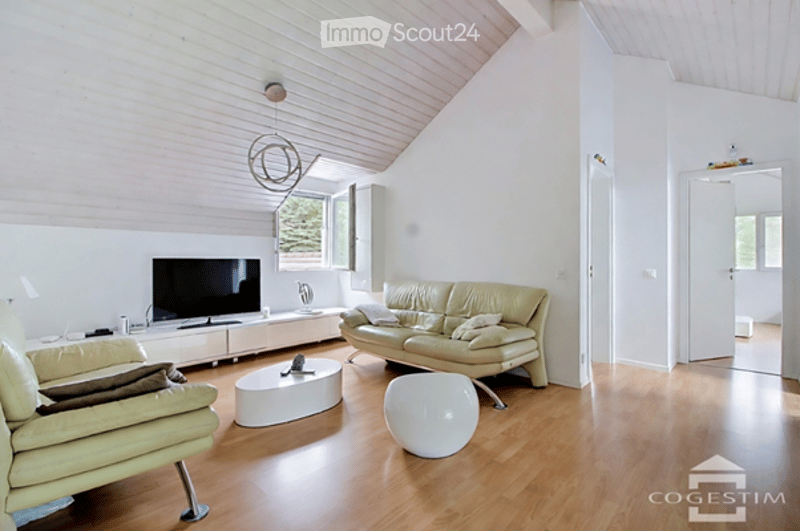 Bright living room with white walls, wooden flooring, and a slanted ceiling. Features a cream-colored sofa, a white coffee table, a television on a stand, and a modern floor lamp.