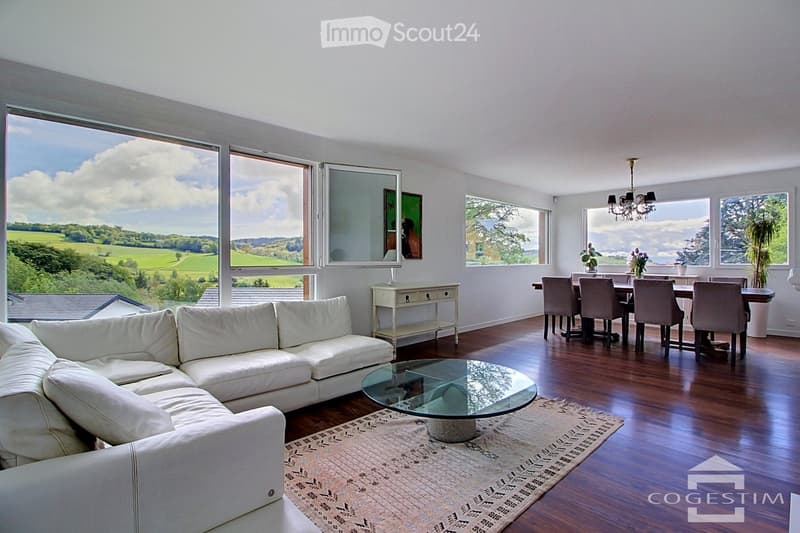 Bright living and dining area with large windows offering scenic views, featuring a white sectional sofa, glass coffee table, dining table with chairs, and a chandelier.