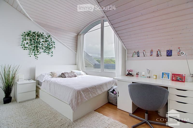 Cozy bedroom with a large bed, nightstand, desk, and chair. The room features wooden flooring, white walls, and large windows with curtains. Decor includes a plant, picture frames, and a small lamp on the desk.