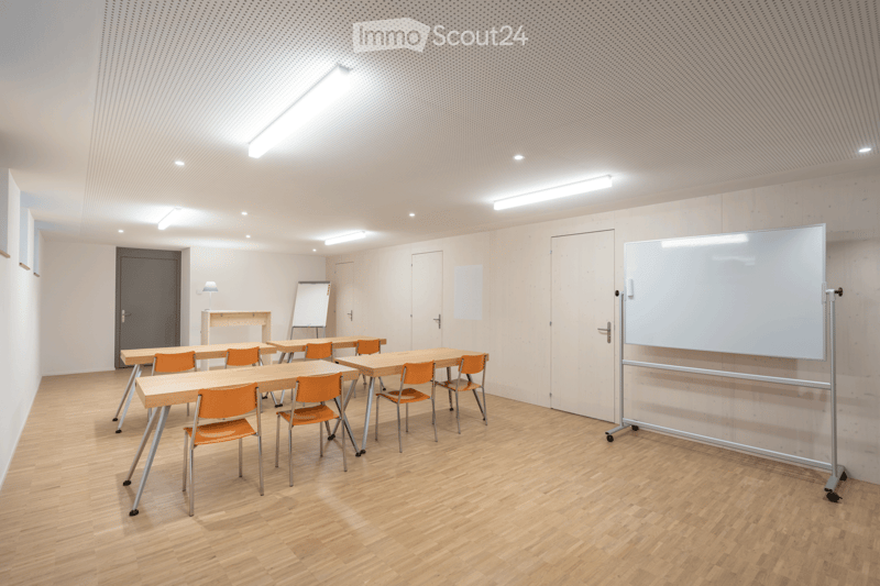 Bright, modern room with wooden flooring, multiple tables and chairs arranged in rows, a large whiteboard, and a projector screen on wheels.