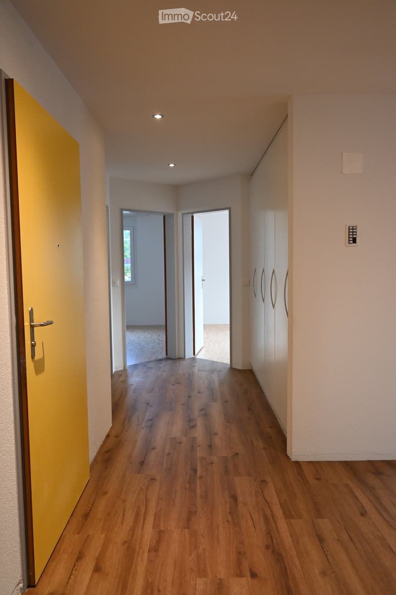 An empty corridor leading to the entrance, with a yellow door and an empty hallway with two open doors, and a wooden floor. There are two walls, one of which has white shelves, while the other is white.