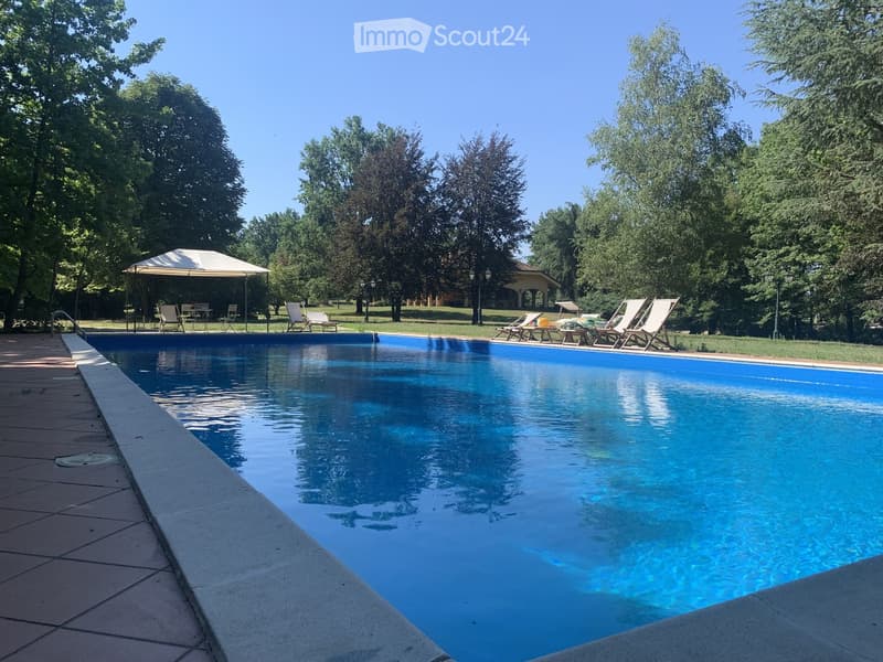 A large swimming pool surrounded by lounge chairs, a gazebo, and trees in a park-like setting.