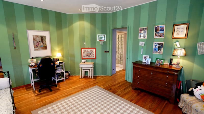 Cozy bedroom with green striped walls, wooden flooring, a bed with a patterned bedspread, a desk with a chair, a dresser with lamps, and several picture frames on the walls.