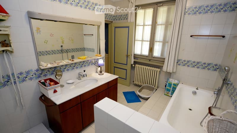 Bathroom with white tiles, a large mirror, a sink with a cabinet, a bathtub, a cat litter box, and a small lamp on the sink counter.