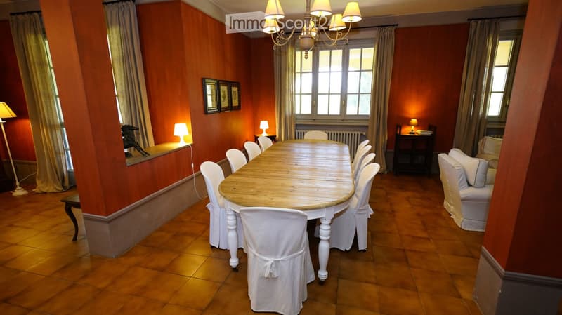 Dining room with a long wooden dining table surrounded by white chairs, a white sofa, table lamps, a chandelier, and picture frames on the wall.
