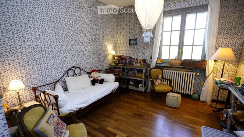 Cozy bedroom with a sofa bed, armchair, small table, lamps, books, toys, and baskets. The room features patterned wallpaper and wooden flooring.