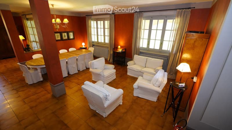 A spacious, well-lit living and dining area with wooden flooring, white furniture including a sofa, armchairs, and a dining table with chairs. The room features large windows with curtains, wall-mounted lamps, and a chandelier.