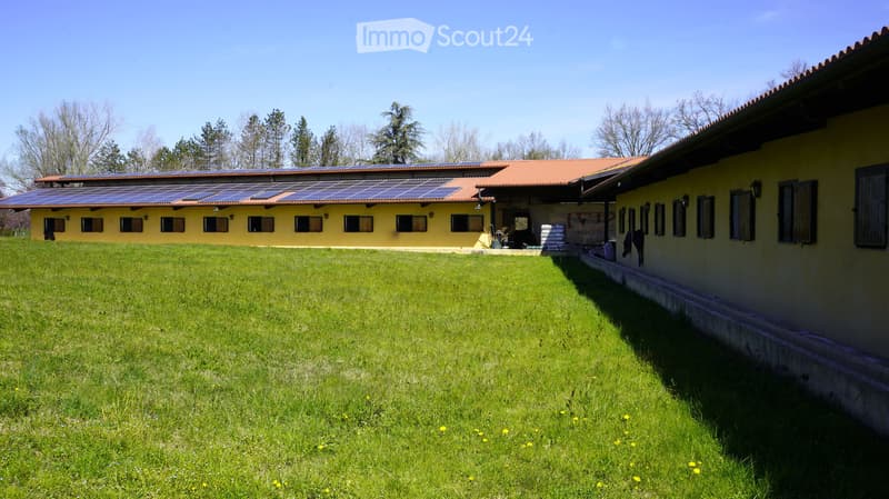 A large, well-maintained lawn in front of a yellow building with a red-tiled roof and solar panels on the roof. The building has multiple windows and a covered entrance area.