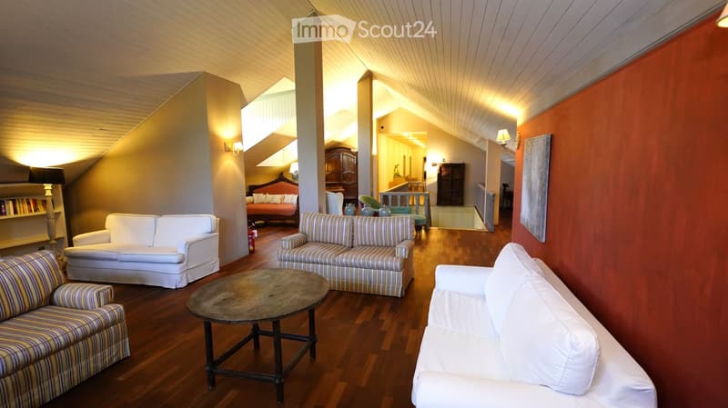 A spacious living room with wooden flooring, white and striped sofas, a coffee table, and a bookshelf. The room features warm lighting and an open layout with a view into another room.