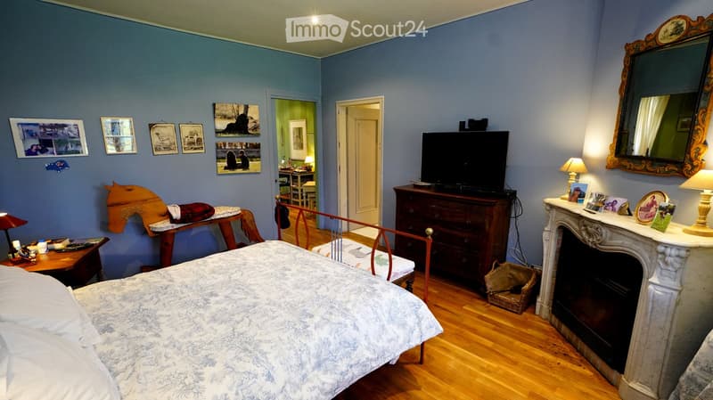 Cozy bedroom with a wooden bed, nightstand, lamp, mirror, television, dressers, fireplace, and a small table with a chair.