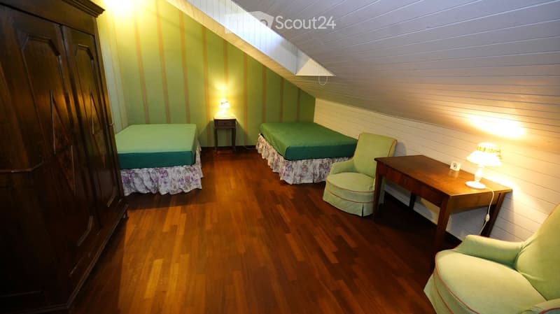Cozy attic bedroom with two single beds, a small table, a chair, and a wardrobe. The room features wooden flooring and slanted ceilings with exposed beams.