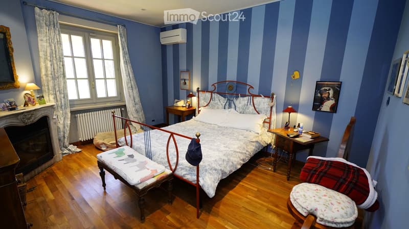 Cozy bedroom with wooden floors, blue striped walls, a metal-framed bed, nightstands, a fireplace, and a window with curtains.