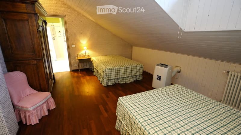 Cozy attic bedroom with wooden floors, two single beds with checkered bedspreads, a pink upholstered chair, a small table with a lamp, and an air conditioner unit.