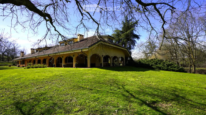 A large, two-story yellow house with a brown tiled roof, surrounded by a well-maintained green lawn and trees.