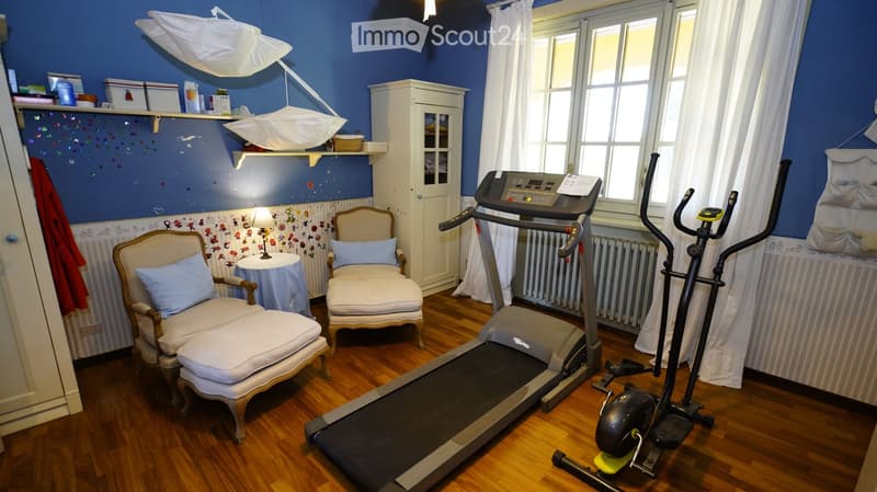 A well-lit room with wooden flooring, featuring a treadmill, exercise bike, two armchairs, a small table, a cabinet, and shelves with various items.