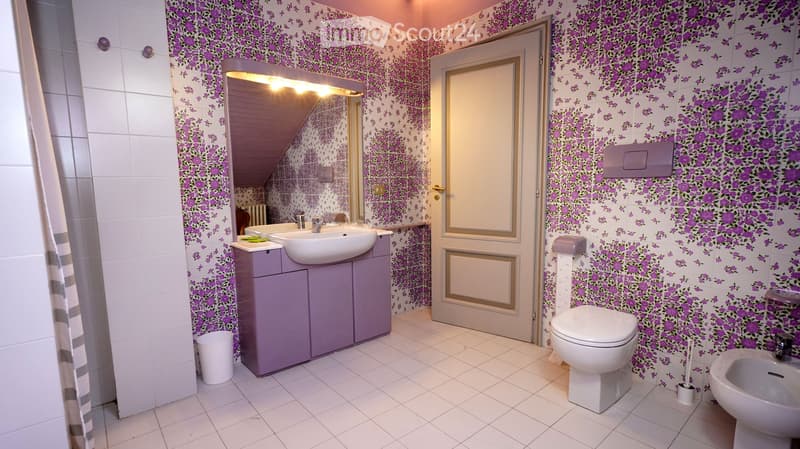 Bathroom with floral wallpaper, white tiles, a purple cabinet with a sink, a toilet, a bidet, and a mirror with built-in lighting.