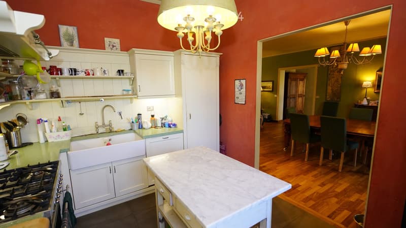 The image shows a cozy kitchen with white cabinets, a green countertop, and a marble island. A sink with a faucet is visible, along with a stove on the left. The kitchen is connected to a dining area with a wooden table and green chairs.