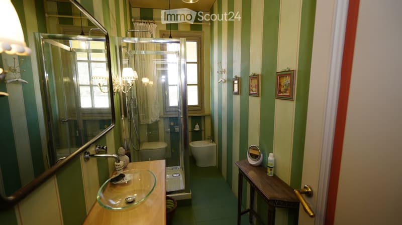 Bathroom with green and white striped walls, a glass shower enclosure, a sink with a mirror, a small table with toiletries, and a toilet and bathtub in the background.