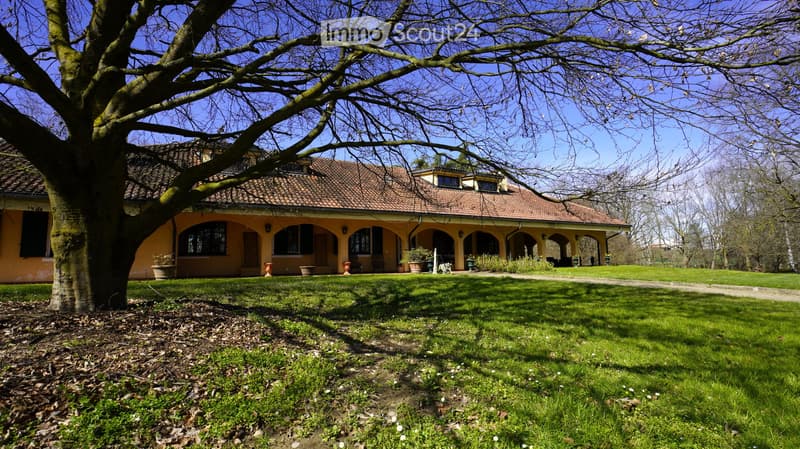 A two-story house with a tiled roof, arched windows, and a covered porch area. The house is surrounded by a well-maintained lawn and trees.