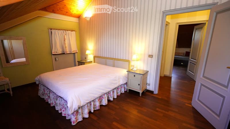 Cozy attic bedroom with wooden floors, a double bed with floral bed skirt, nightstands with lamps, a mirror, and an open door leading to another room.