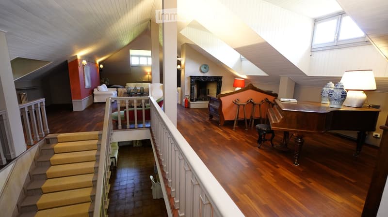 Spacious attic room with wooden floors, slanted ceilings, fireplace, piano, and seating area.
