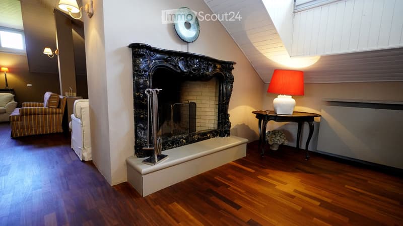 Living room with a large ornate fireplace, wooden flooring, a sofa, armchairs, a small table with a lamp, and a decorative mirror on the wall.