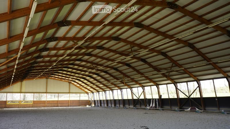 Large, open indoor space with a high wooden truss ceiling and exposed beams, featuring a sandy or gravel floor and large windows along one side.