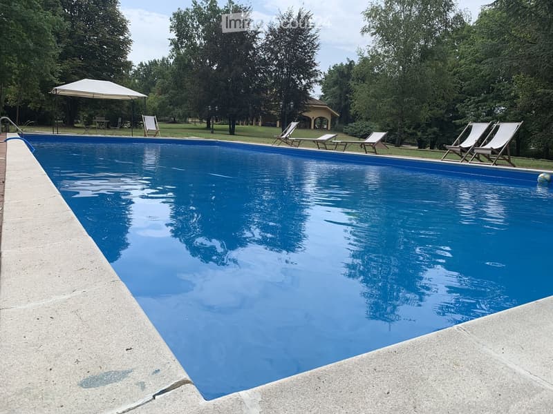 A large swimming pool with clear blue water, surrounded by lounge chairs and a small gazebo in a park-like setting with trees and greenery.