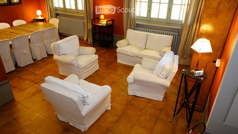 Living room with wooden flooring, white sofa, armchairs, a small table, and lamps. The room has large windows with curtains and a dining area with a table and chairs.