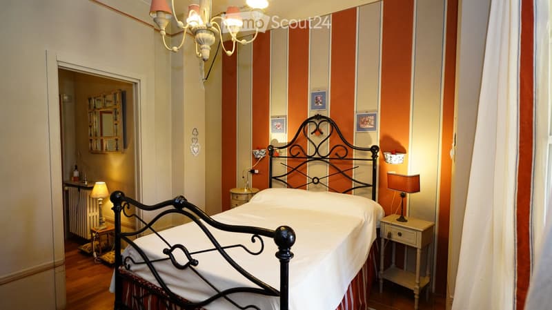 Cozy bedroom with a black metal bed, white bedding, two nightstands with lamps, a mirror, and a radiator. The walls are painted in a striped orange and cream pattern, and the floor is wooden.