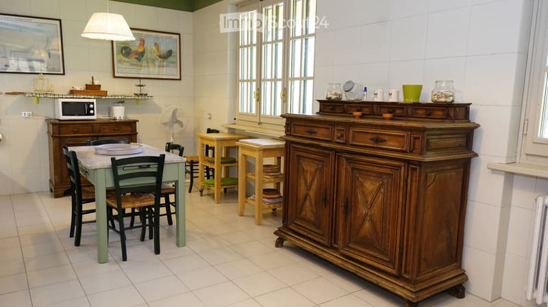 Bright kitchen with white tiled walls and floors, featuring a wooden cabinet, a green table with chairs, a microwave, and a small fan. There are also decorative items on the shelves and a small wooden stool.