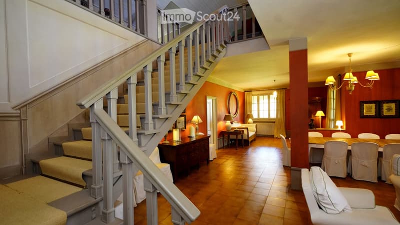 Spacious, well-lit living area with wooden flooring, a staircase leading to an upper level, a seating area with a sofa and chairs, a dining table with chairs, and decorative lamps.