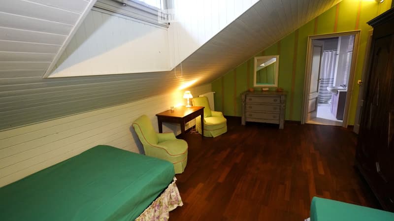 Cozy attic bedroom with wooden floors, slanted ceilings, green walls, a bed with green bedding, a small table with a lamp, two green armchairs, and a dresser. A bathroom is visible through an open doorway.