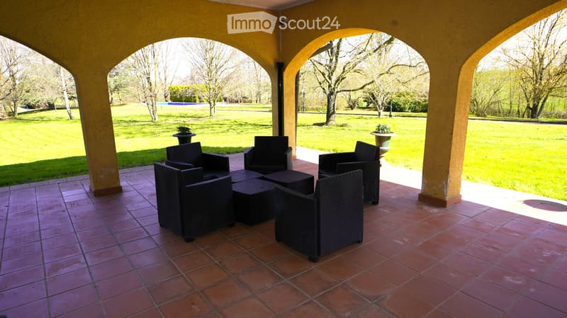 Arched outdoor patio with tiled floor, black wicker chairs and tables, overlooking a grassy park area.