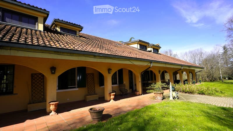 Two-story house with a tiled roof, arched covered walkway, and well-maintained garden with potted plants and a lawn.