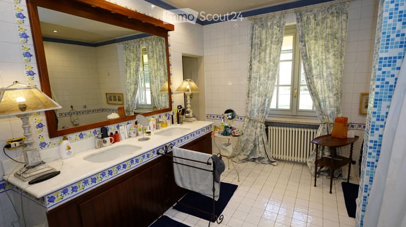 Bathroom with double sink, large mirror, floral tile border, two lamps, towel rack, small table, and various toiletries.