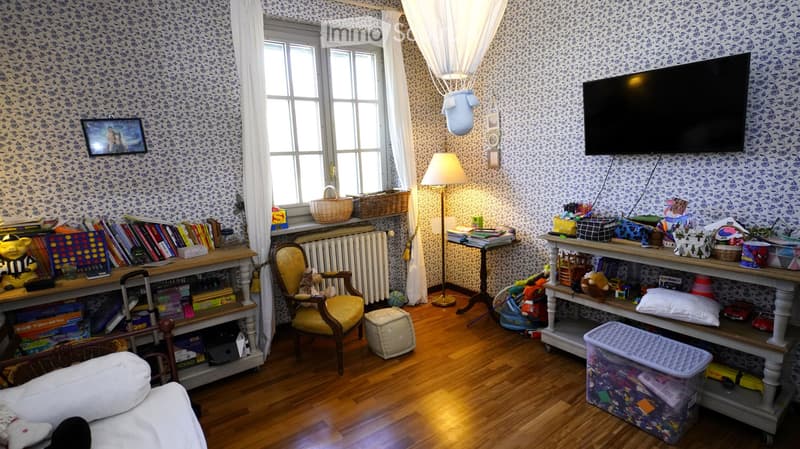A cozy, well-lit room with wooden flooring, patterned wallpaper, and large windows. Features a sofa, armchair, bookshelf, floor lamp, television, and various toys and storage bins.