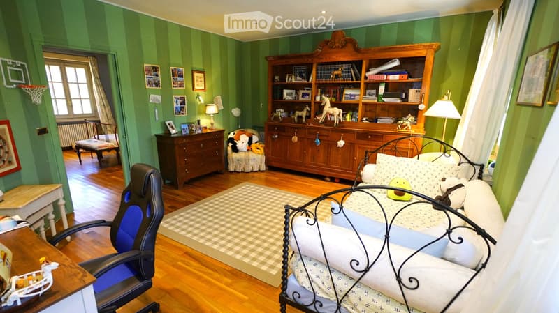 A well-furnished bedroom with green striped walls, wooden flooring, a metal-framed bed, a wooden cabinet, a desk chair, and decorative items including toys and picture frames.