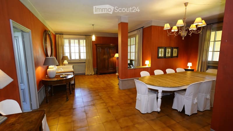 Spacious dining area with wooden floors, warm orange walls, large windows with curtains, a round wooden dining table with white chairs, a side table with a lamp, and a wooden cabinet.