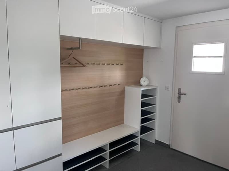 Empty room with white cabinets, wooden wall paneling, built-in bench, and a small white shelf unit with a clock on top.