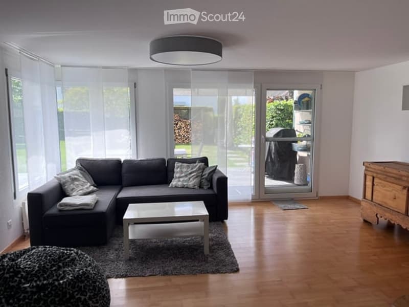 Modern living room with large windows, grey sectional sofa, coffee table, ottoman, and floor lamp.