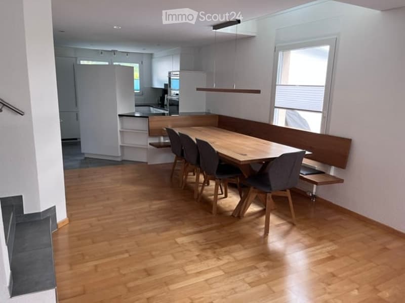 Bright dining area with a wooden dining table surrounded by modern chairs and a built-in bench, connected to a modern kitchen with white cabinets and stainless steel appliances.