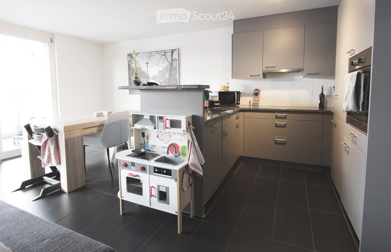 Modern kitchen with grey cabinets, black tiled floor, and a small wooden table with a white chair. A toy stove is placed near the sink, and a microwave is on the counter.