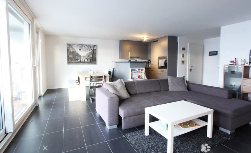 Modern living and dining area with a grey sectional sofa, white coffee table, dining table with chairs, and an open kitchen with cabinets, microwave, oven, and refrigerator.