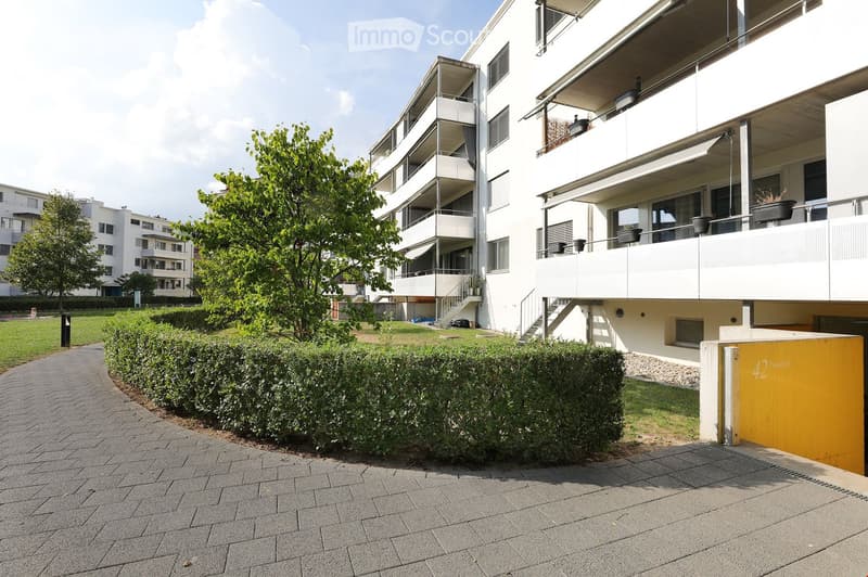 Modern multi-story residential building with balconies, surrounded by greenery and a paved walkway.