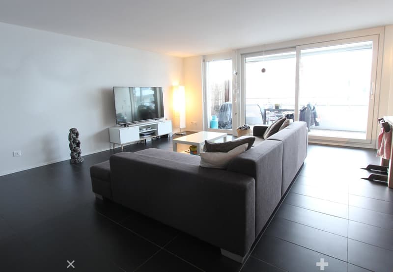 Modern living room with dark tiled flooring, grey sectional sofa, coffee table, flat-screen TV on a white stand, and a decorative sculpture on the floor.