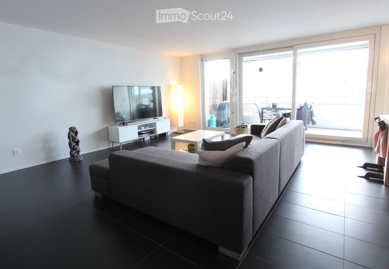 Modern living room with dark tiled flooring, grey sectional sofa, coffee table, flat-screen TV on a white stand, and a decorative sculpture on the floor.