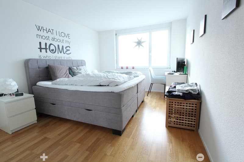Bright bedroom with wooden flooring, a large bed with grey bedding, a nightstand with a lamp, a desk with a computer, and a chair. A decorative wall quote is visible on the wall.