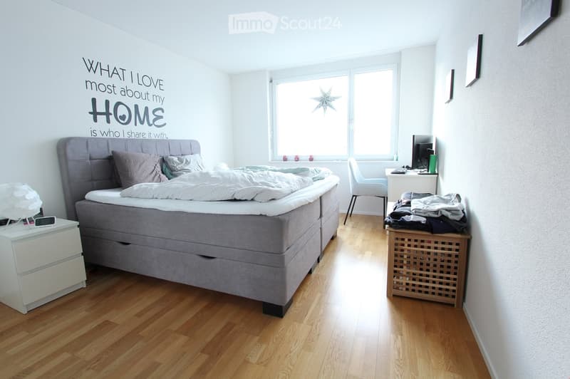 Bright bedroom with wooden flooring, a large bed with grey bedding, a nightstand with a lamp, a desk with a computer, and a chair. A decorative wall quote is visible on the wall.